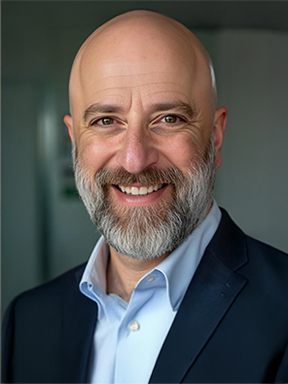A man with a salt-and-pepper beard wearing a blue dress shirt and dark blue suit coat.