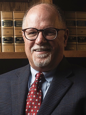 A smiling man with gray facial hair wearing glasses, a blue suit and a red necktie.