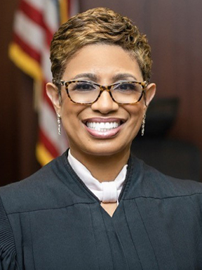 A woman with short hair and glasses wearing a judge's robe with an American flag in the background.