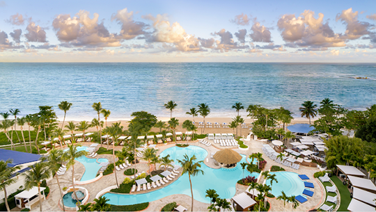 View of hotel pools looking out to the ocean under a blue sky with pink puffy clouds.