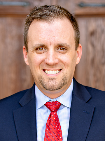 Young man with short brown hair wearing a navy blue suit, light blue dress shirt and red patterned necktie.