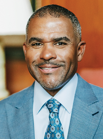 A man in a light blue suite, blue dress shirt and blue flowered necktie smiling warmly at the camera.