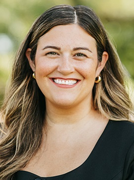 Woman with long brunette hair in a cornflower black suit and gold hoop earrings.