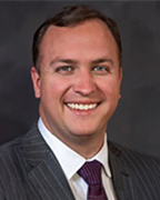 A man in a gray pinstriped suit with a white dress shirt wearing a burgundy necktie.
